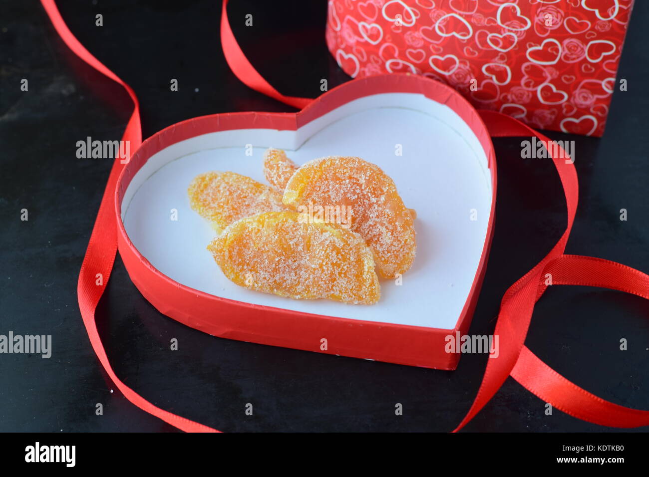Kandierte Orangenscheiben in Herzform rot Deckel aus einer Box mit rotem Band auf einem schwarzen Hintergrund. st. Valentinstag. Romantik, Liebe Stockfoto