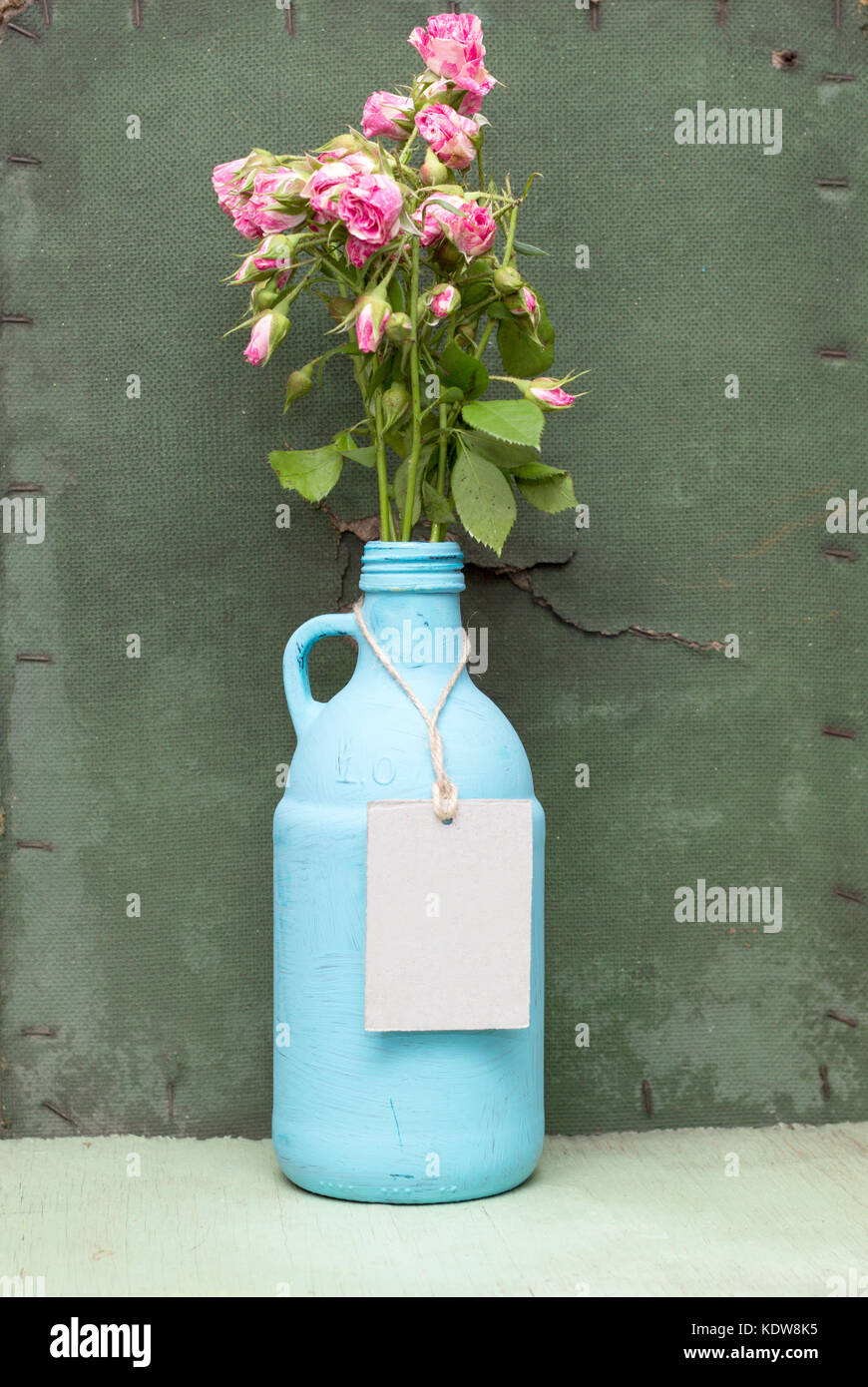 Malte blaue Flasche mit Kraft lable. grunge Grün Holz- Hintergrund. alte Flasche tag Mock-up. blaue Kanne mit rosa Rosen. Stockfoto