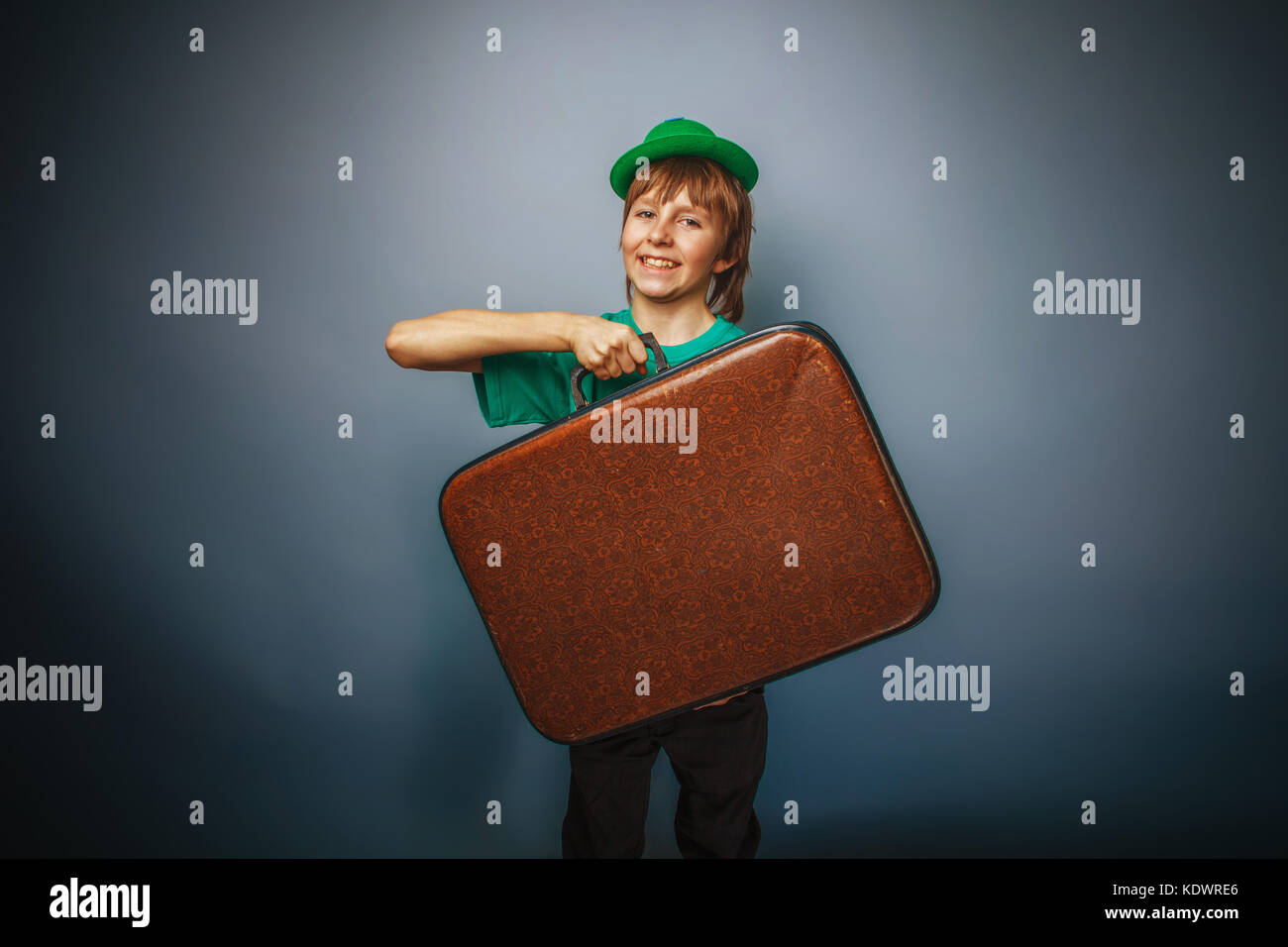 Europäische Erscheinungsbild Teenager Junge in ein Shirt in grünen Hut hält einen alten Koffer in der Hand auf grauem Hintergrund, Abenteuer cross Prozess Stockfoto