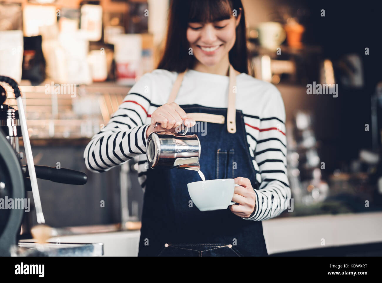 Asien Frau barista gießen Sie Milch in die heiße Tasse Kaffee an der Bar vor der Maschine im Cafe Restaurant, Essen Geschäftsinhaber Konzept Stockfoto