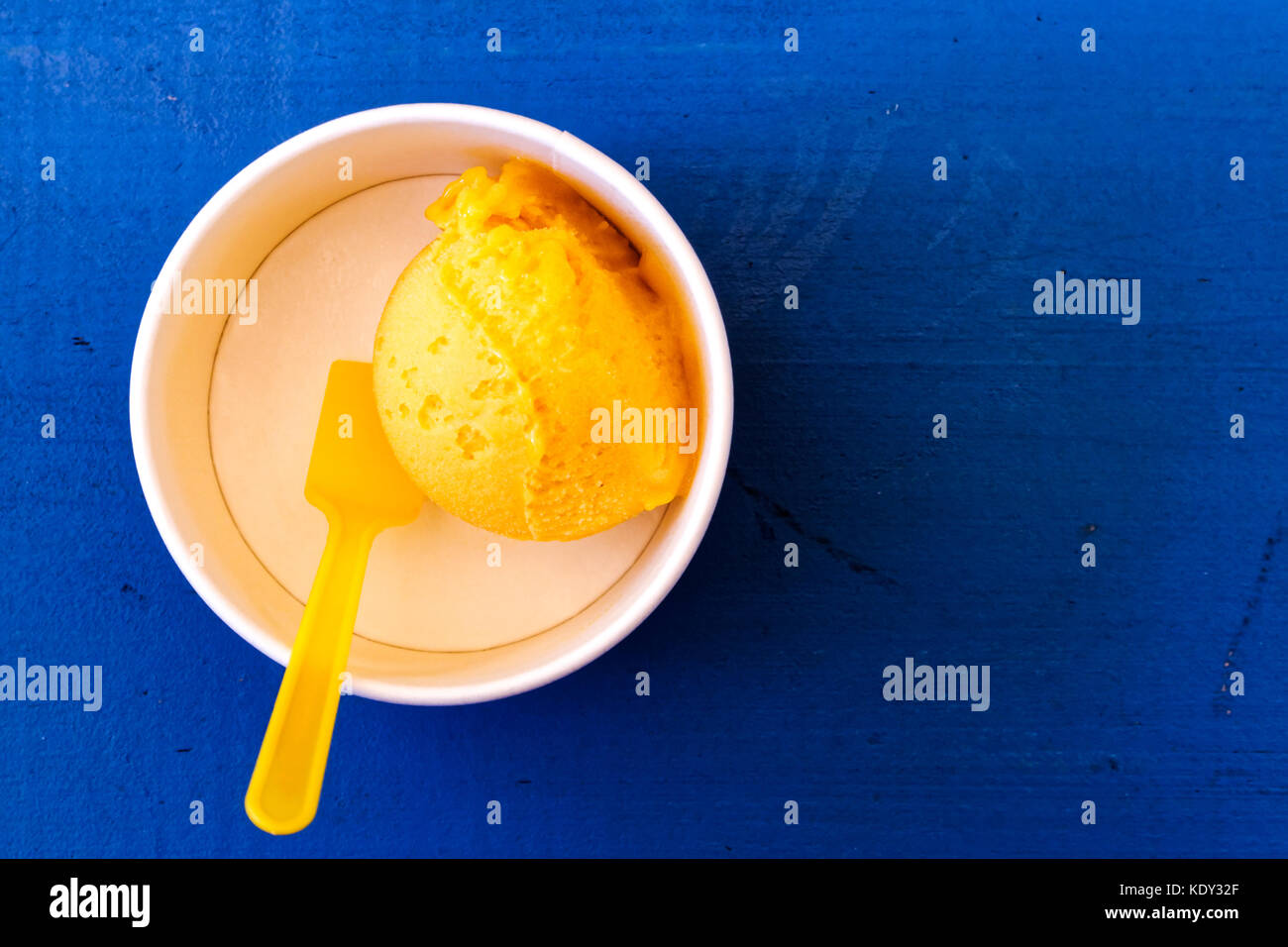 Schaufel von Mango Eis in Pappbecher mit plastiklöffel auf Blau lackiertes Holz von oben isoliert. Stockfoto