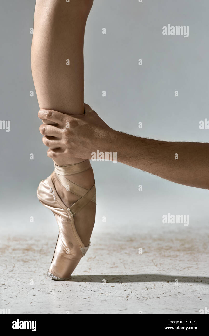 Die Hand des Mannes hält das Bein des Ballerina auf dem grauen Hintergrund im Studio. Mädchen trägt beige Spitzenschuhe und steht auf der Zehe. closeup. Vertikale Stockfoto