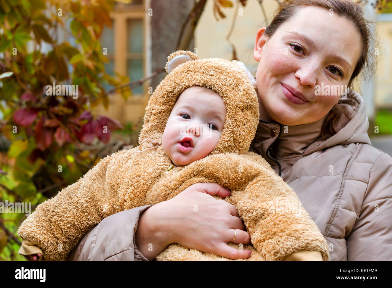 Frau mit Baby in den Armen, auf dem Hintergrund der Herbst Park posieren. Kind gekleidet in warmen stilisierte Teddy - Bär Kostüm, Spaß am Arm der Mutter. Familie w Stockfoto