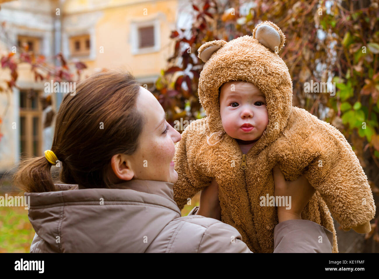 Frau mit Baby in den Armen, auf dem Hintergrund der Herbst Park posieren. Kind gekleidet in warmen stilisierte Teddy - Bär Kostüm, Spaß am Arm der Mutter. Familie w Stockfoto