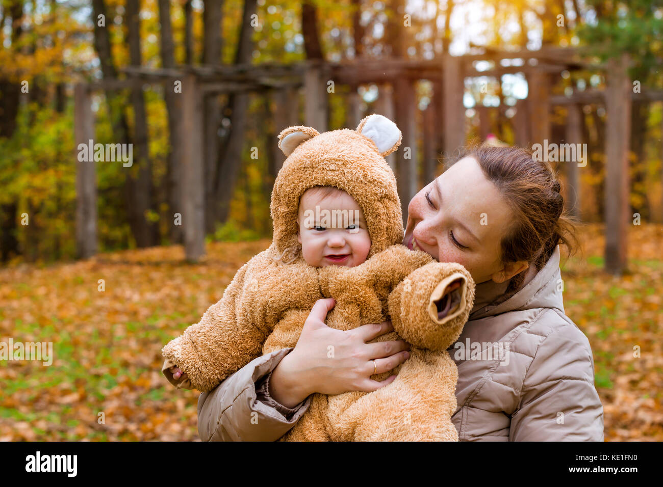 Frau mit Baby in den Armen, auf dem Hintergrund der Herbst Park posieren. Kind gekleidet in warmen stilisierte Teddy - Bär Kostüm, Spaß am Arm der Mutter. Familie w Stockfoto