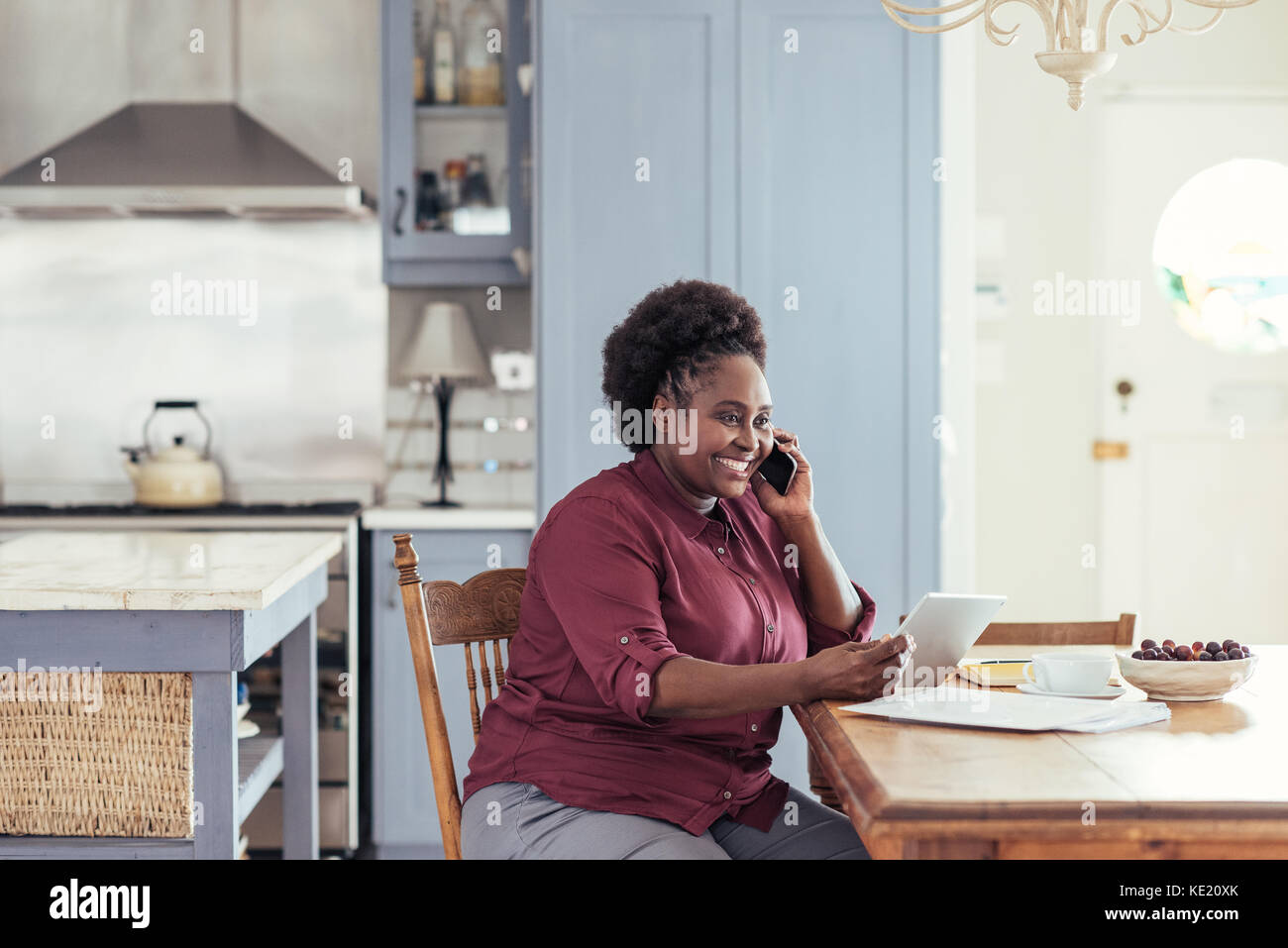 Lächelnde junge afrikanische Frau mit einem digitalen Tablet und Gespräche am Mobiltelefon, während an ihrem Küchentisch sitzen von zu Hause aus arbeiten Stockfoto
