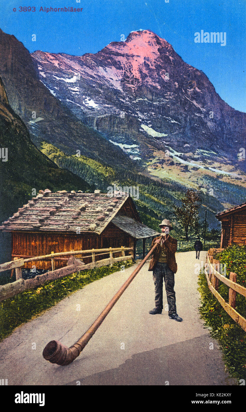 Mann spielt alpine Horn. Alpine Szene mit Bergen im Hintergrund. Holz- Hütte/Chalet. Sehr lange Wind instrument Stockfoto