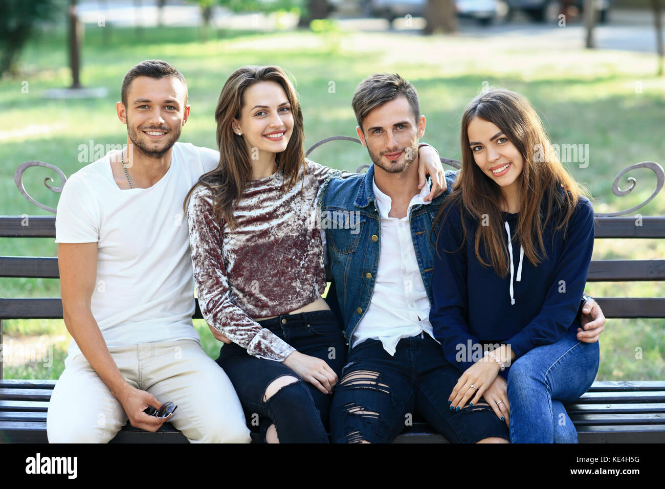 Vier Frauen und Männern sitzt auf der Bank in der Umarmung. Gerne stilvolle junge Freunde auf der Bank draußen Posing, lächelnd und mit Blick auf die Kamera. schönen, gesunden Stockfoto
