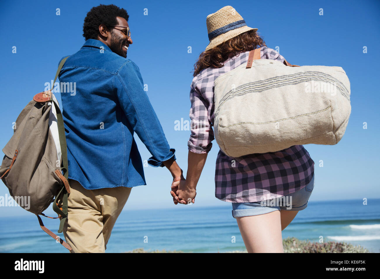 Multiethnisches Paar, das sich die Hände hält und am sonnigen Sommerstrand am Ozean spazierengeht Stockfoto