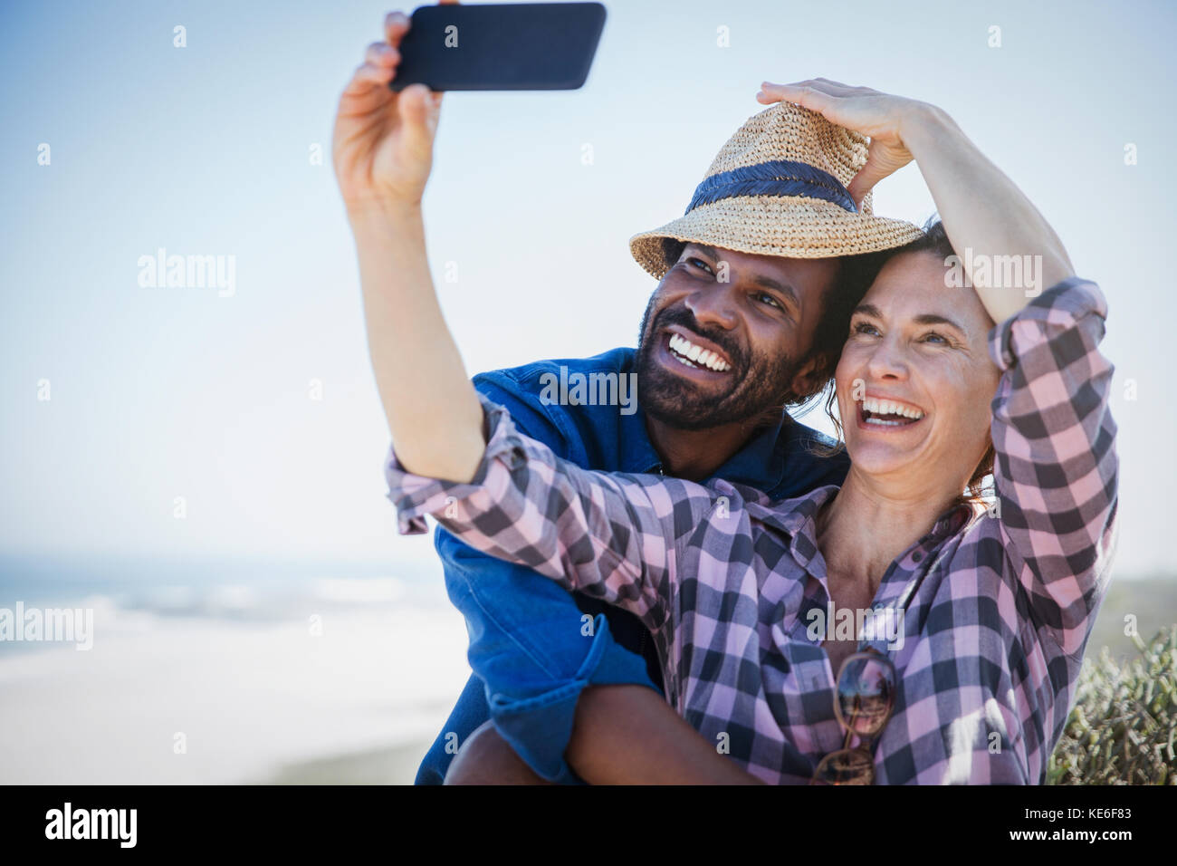 Verspieltes, lächelndes multiethnisches Paar, das Selfie mit dem Kameratelefon am sonnigen Sommerstrand macht Stockfoto
