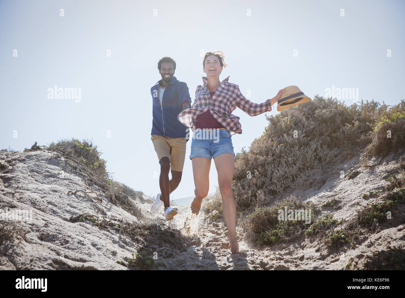 Spielerisches, energiegeladenes multiethnisches Paar, das auf einem sonnigen Sandstrandabschnitt im Sommer läuft Stockfoto