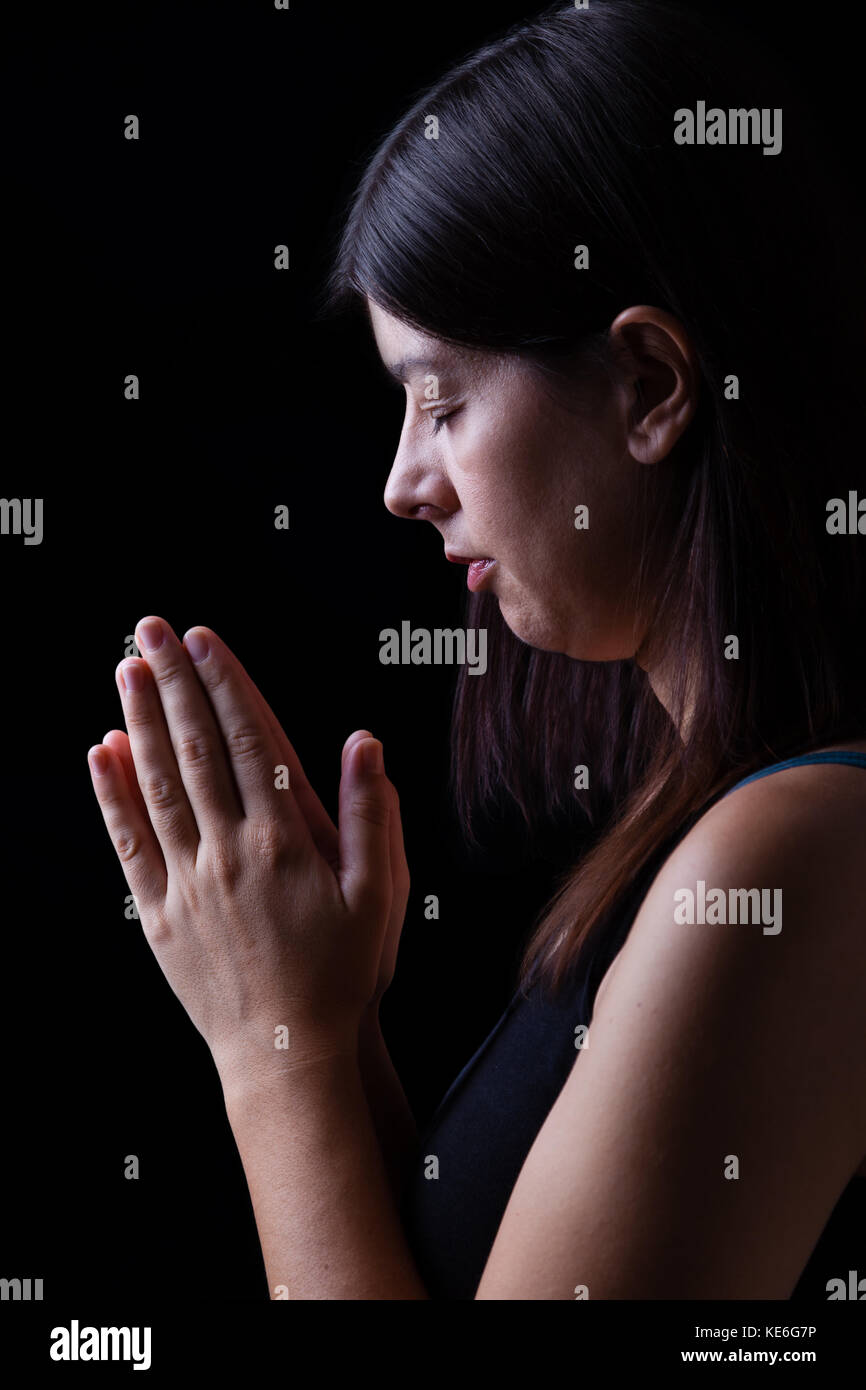 Treue Frau beten, mit Händen in Anbetung zu Gott gefaltet, Kopf nach unten, die Augen geschlossen, in religiöser Inbrunst. schwarzer Hintergrund beten Gebet Handflächen zusammen Stockfoto