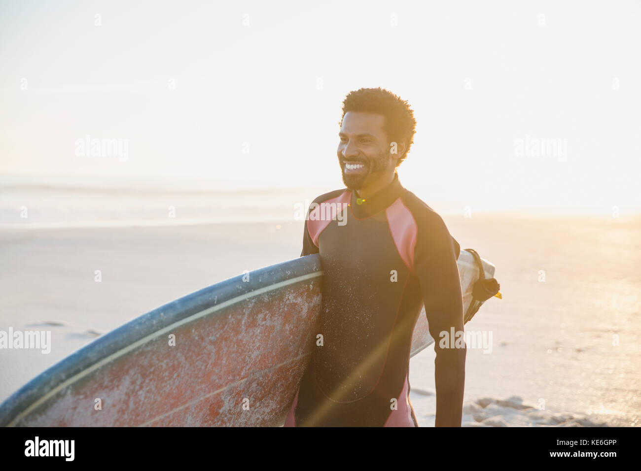 Lächelnder männlicher Surfer, der mit Surfbrett am sonnigen Sommerstrand spazierengeht Stockfoto