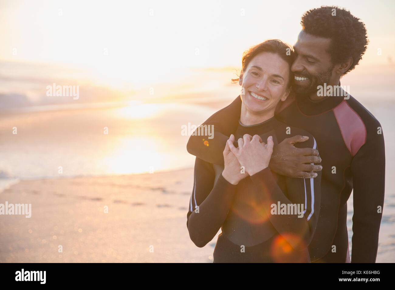 Lächelndes, liebevolles multiethnisches Paar in nassen Anzügen am sonnigen Strand bei Sonnenuntergang im Sommer Stockfoto