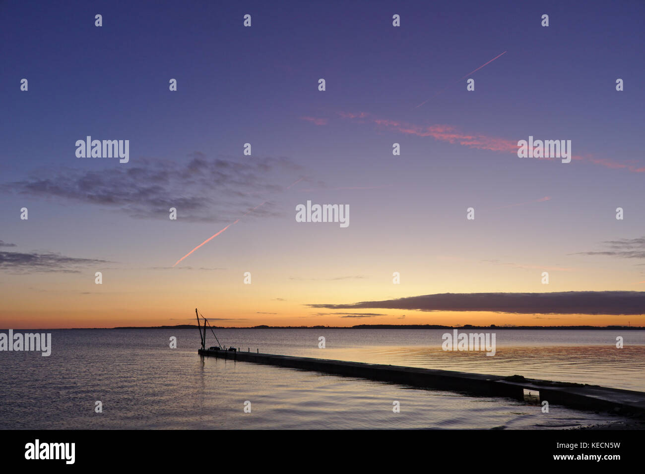 Pier vor Sonnenaufgang mit kleinen Kran gegen den Morgen Himmel und leuchtend roten Kondensstreifen silhouetted Overhead Stockfoto