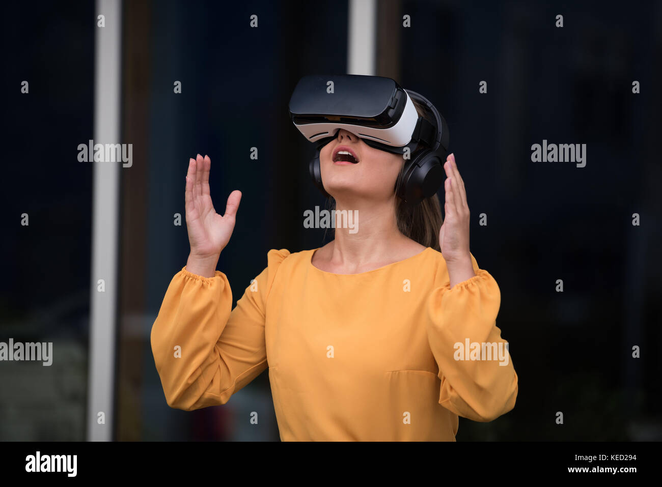Junge lächelnde Geschäftsfrau, tragen gelbe Kleid, virtual reality Brille und Kopfhörer im Freien vor dem Büro Stockfoto