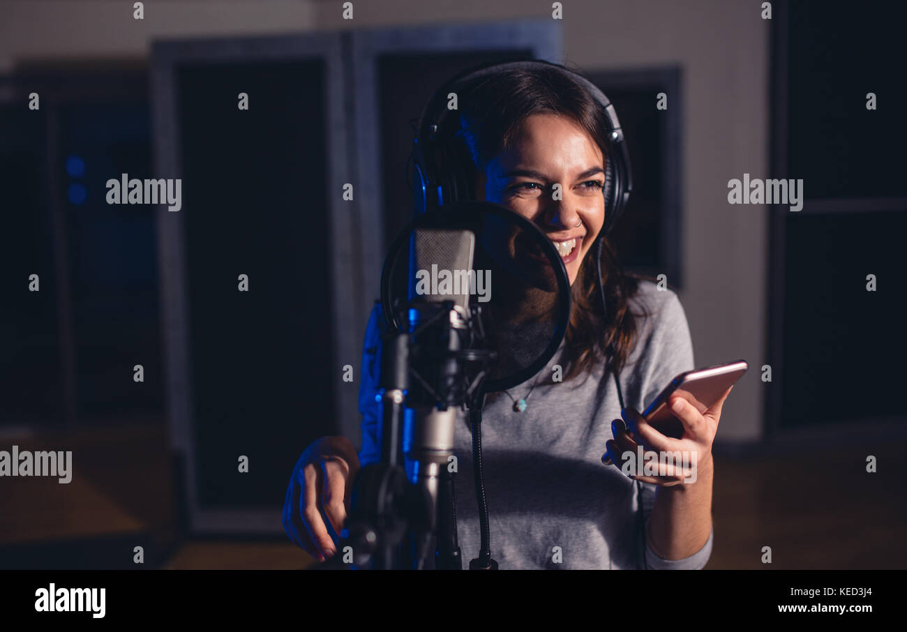 Lächelnd weibliche Wiedergabe Sänger mit Mikrofon und Handy im Aufnahmestudio. Frau mit der Aufnahme eines Songs für Ihr Album im Music Studio. Stockfoto