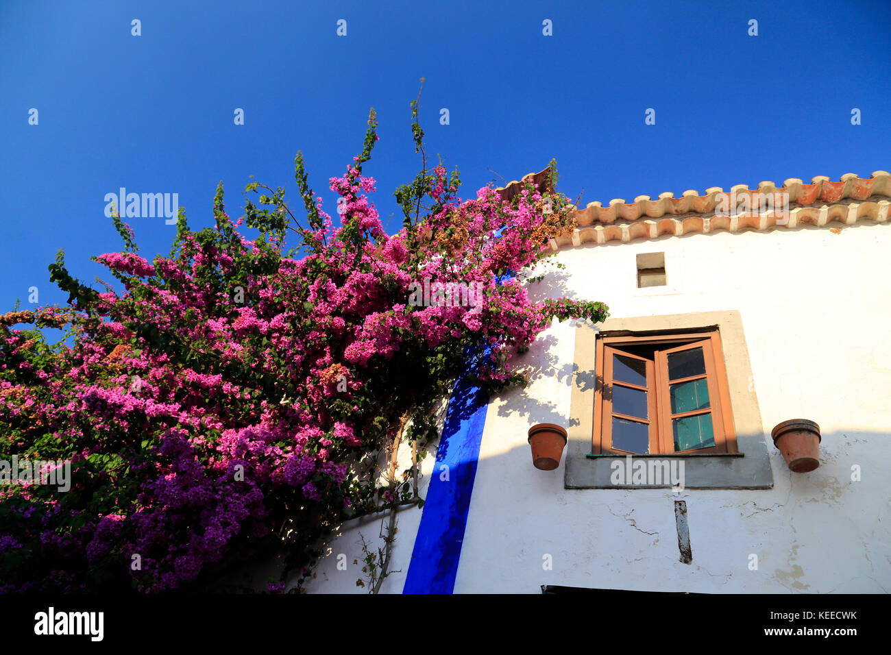 Tief helle rosafarbene Bougainvillea Portugiesischen gegen eine weiße Wand mit gelben Fensterrahmen und Blau umrandete unter pan Ziegeldach und einem intensiven blauen Himmel. Stockfoto