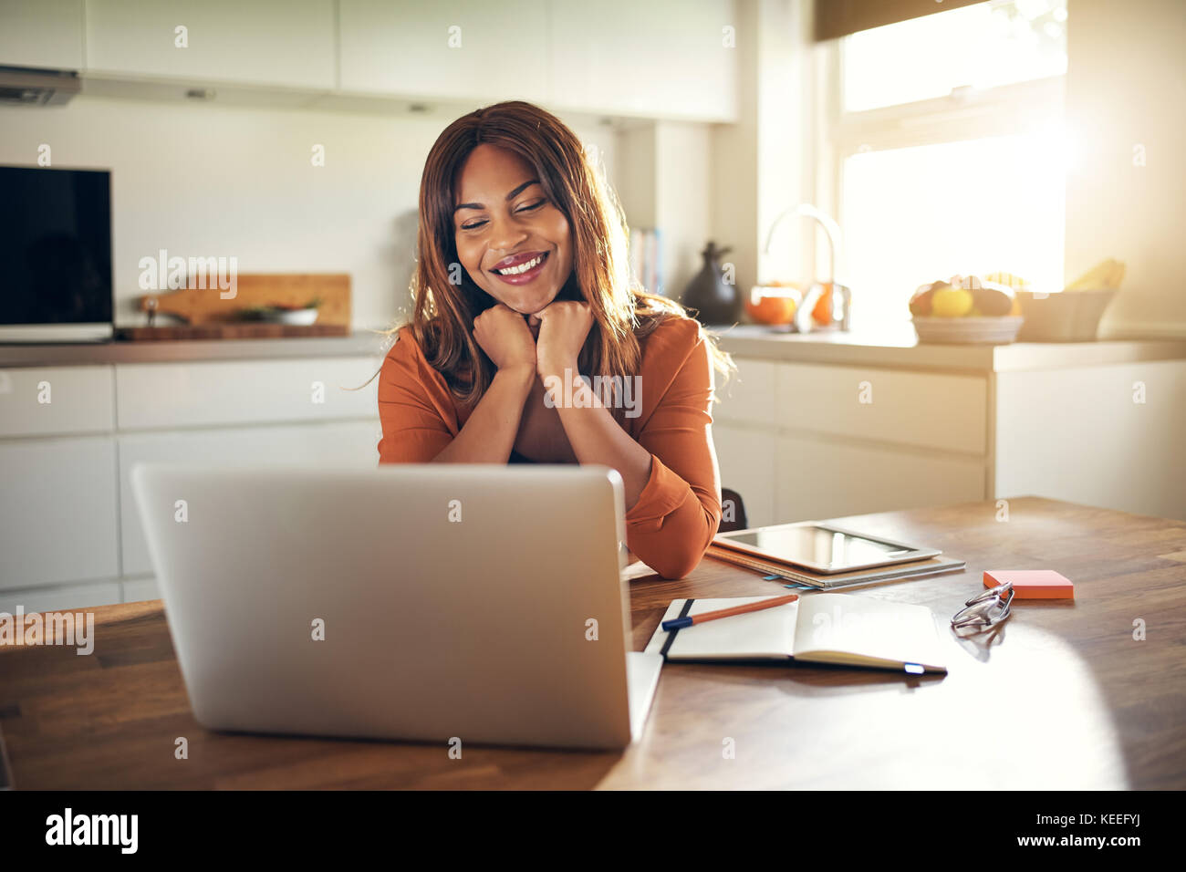 Lächelnden jungen afrikanischen weibliche Unternehmer sitzen an ihrem Küchentisch mit einem Laptop von zu Hause aus arbeiten Stockfoto