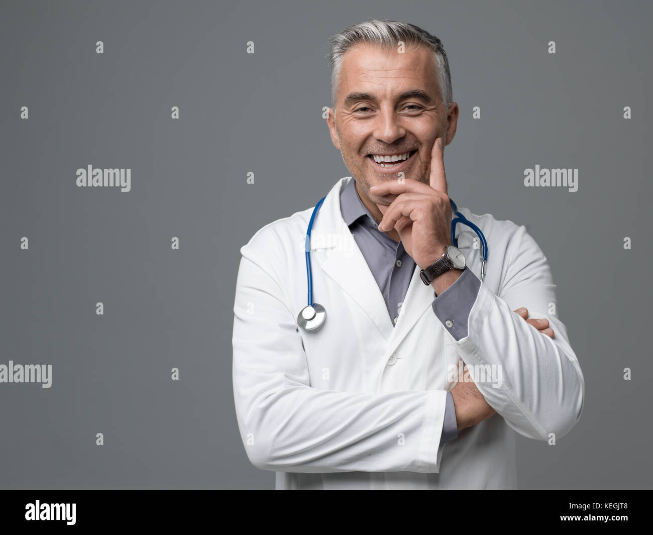 Lächelnd zuversichtlich reife Arzt auf grauem Hintergrund posiert, er blickt auf Kamera Stockfoto