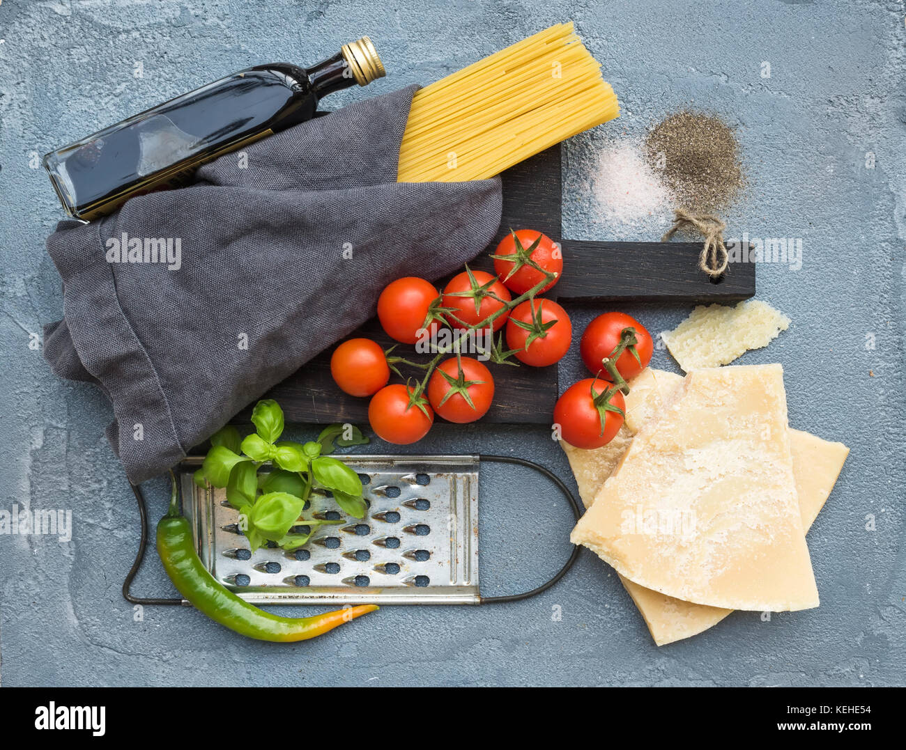 Zutaten für Kochen Pasta. Spaghetti auf dunklen Holzplatte, Parmesan, Tomaten, metallreibebrett, Olivenöl und frischem Basilikum auf Grau concr Stockfoto
