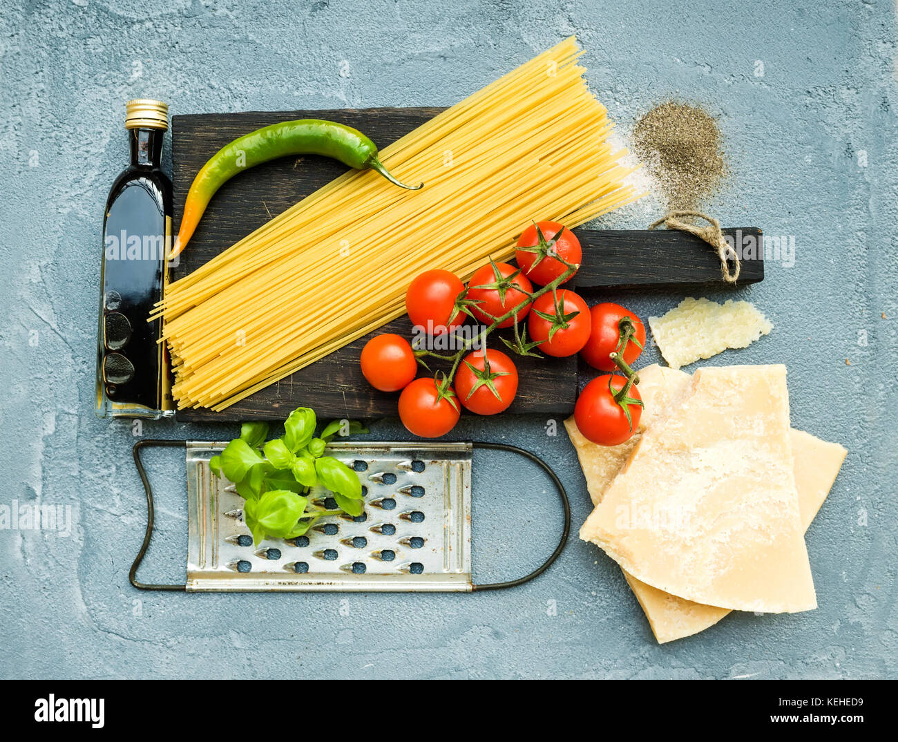Zutaten für Kochen Pasta. Spaghetti auf dunklen Holzplatte, Parmesan, Tomaten, metallreibebrett, Olivenöl und frischem Basilikum auf grau-blau Stockfoto