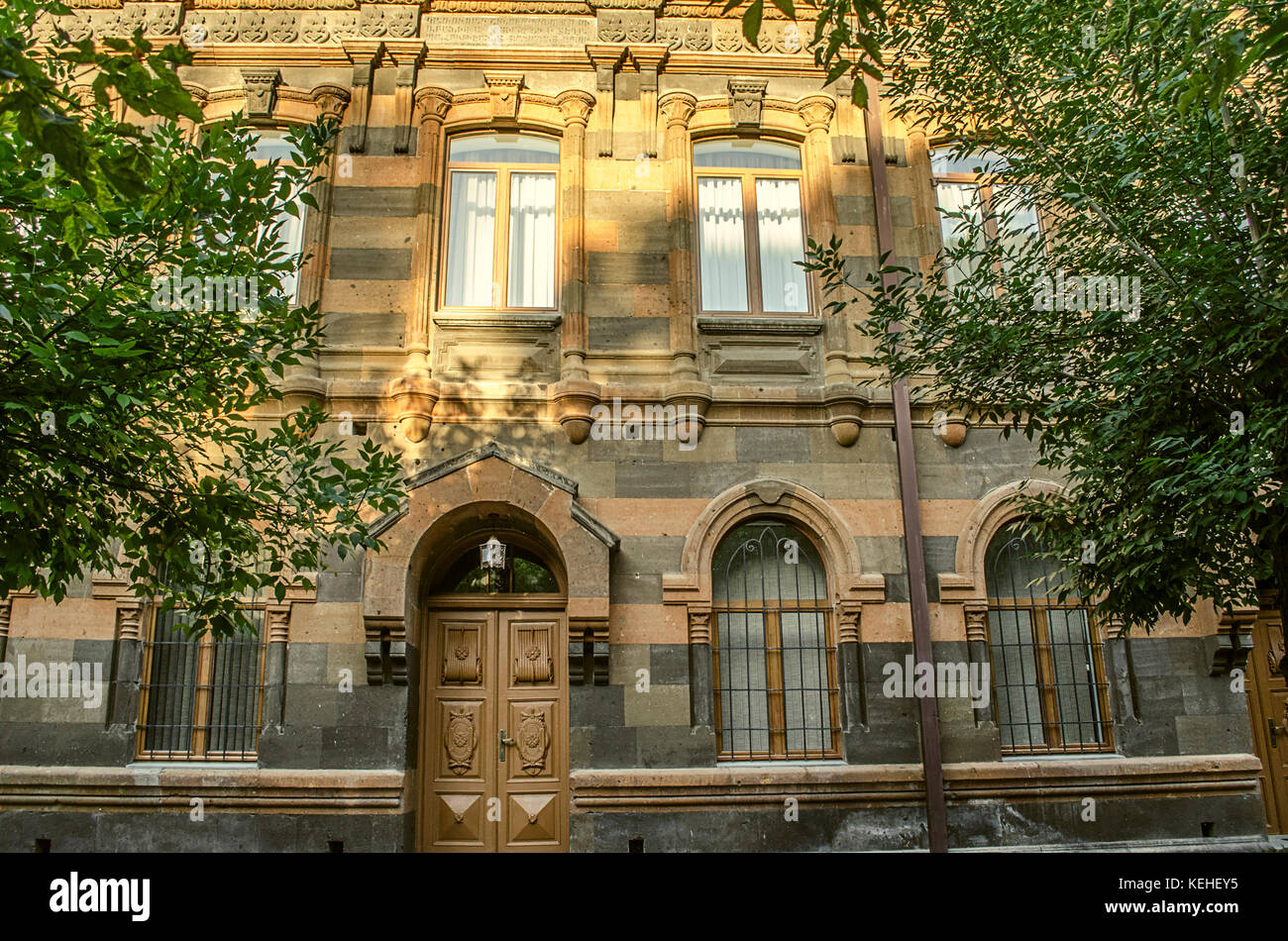 Fassade des alten Gebäudes von mehrfarbigen Tuffstein mit geschnitzten Bögen, Säulen und Simse umgeben von Bäumen Stockfoto