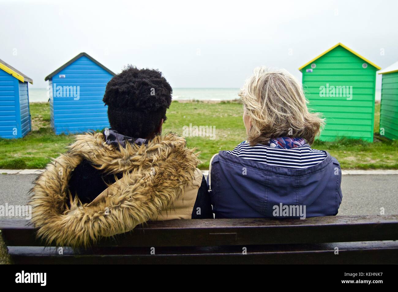 Eine schwarze und eine weiße Dame auf einer Bank vor Strandhütten, Littlehampton UK Stockfoto