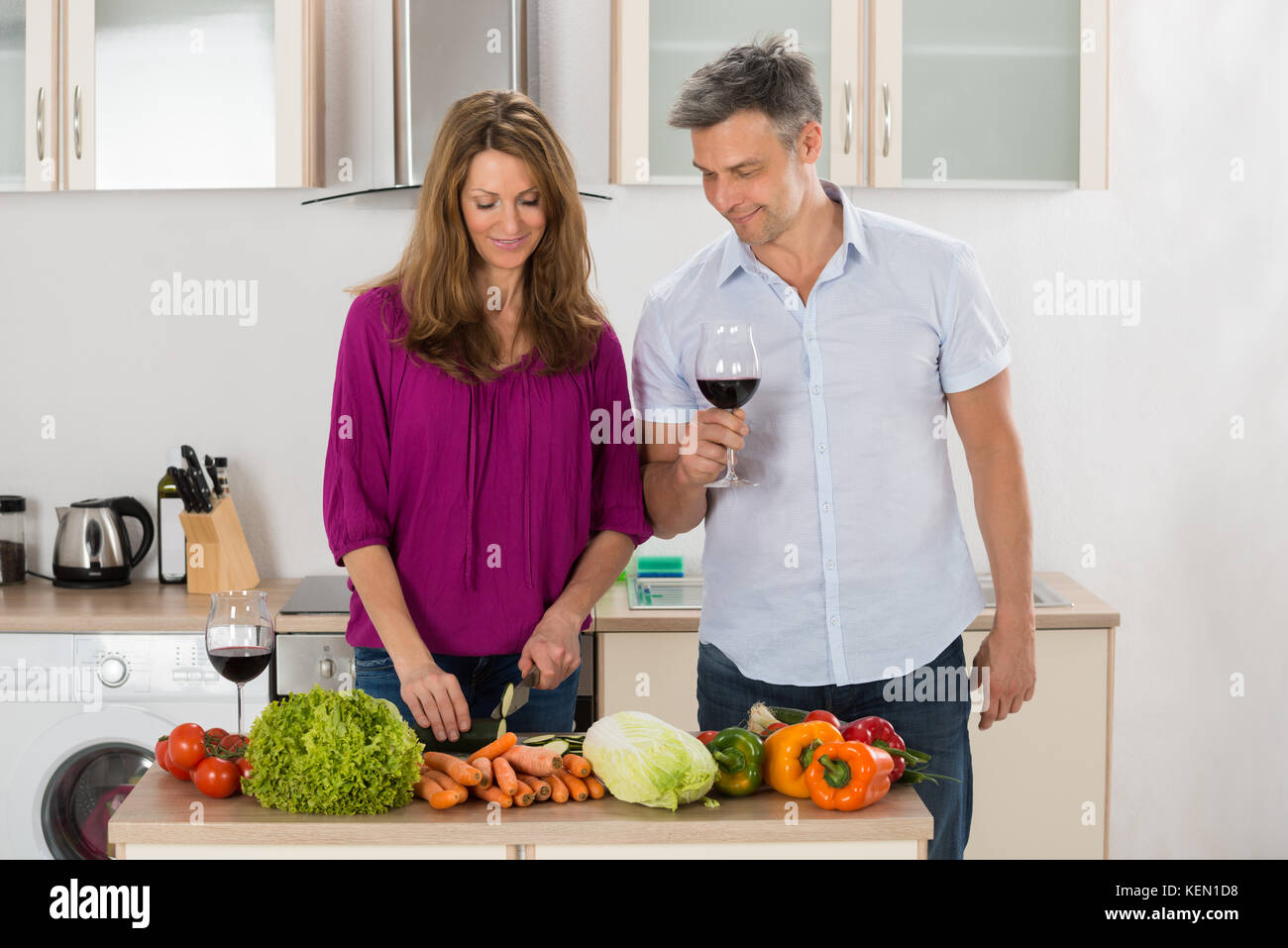 Glückliche Frau Schneiden von Gemüse in der Küche, während ihr Ehemann Holding Glas Wein Stockfoto