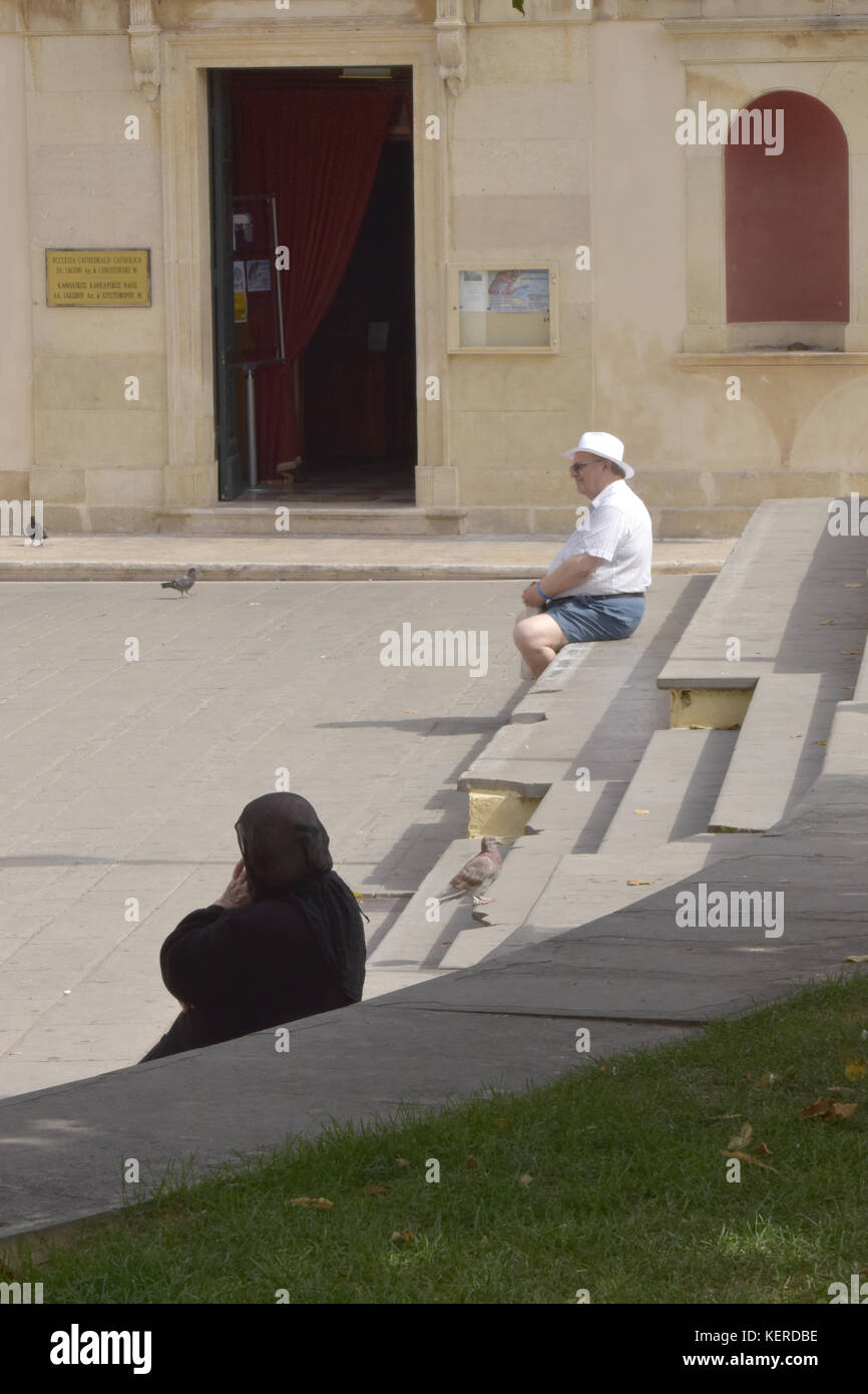 Ein älterer Herr neben einer griechischen Frau oder eine Frau in der traditionellen griechischen Kostüm oder Kleid auf einige Schritte außerhalb einer Kirche in Kerkira, Korfu sitzen. Stockfoto