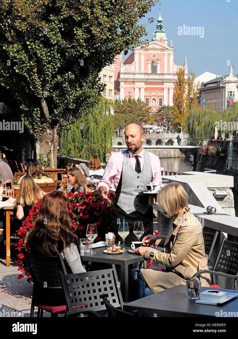 Kellner mit Kunden an Café im Freien entlang der Ljubljanica in der Altstadt von Ljubljana, Slowenien, mit Franziskaner Kirche der Verkündigung in backgroun Stockfoto