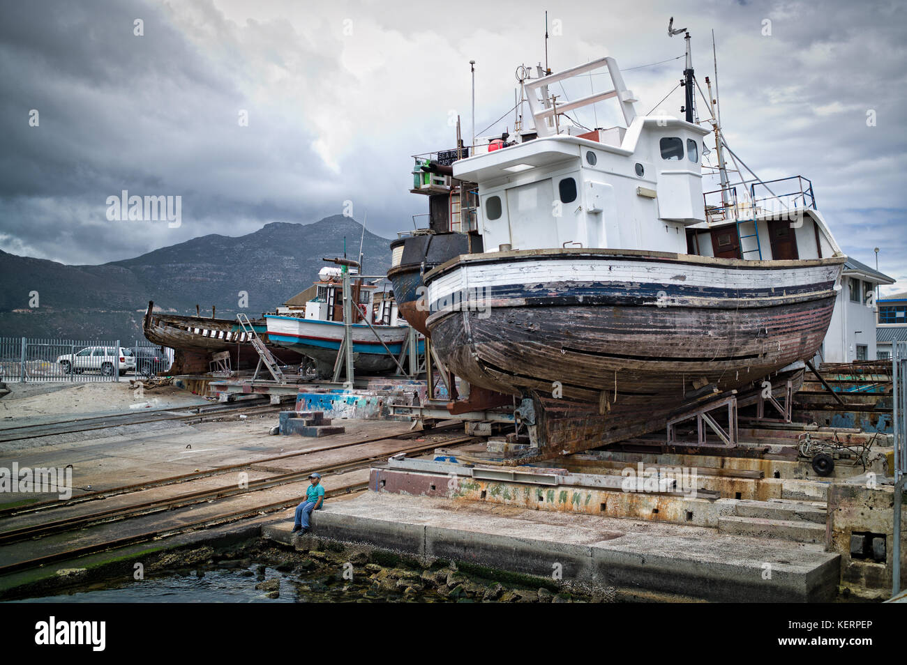 Ein Junge sitzt in der Werft mit Schiffen, die in den Hafen von Hout Bay, Kapstadt, Südafrika repariert werden. Stockfoto