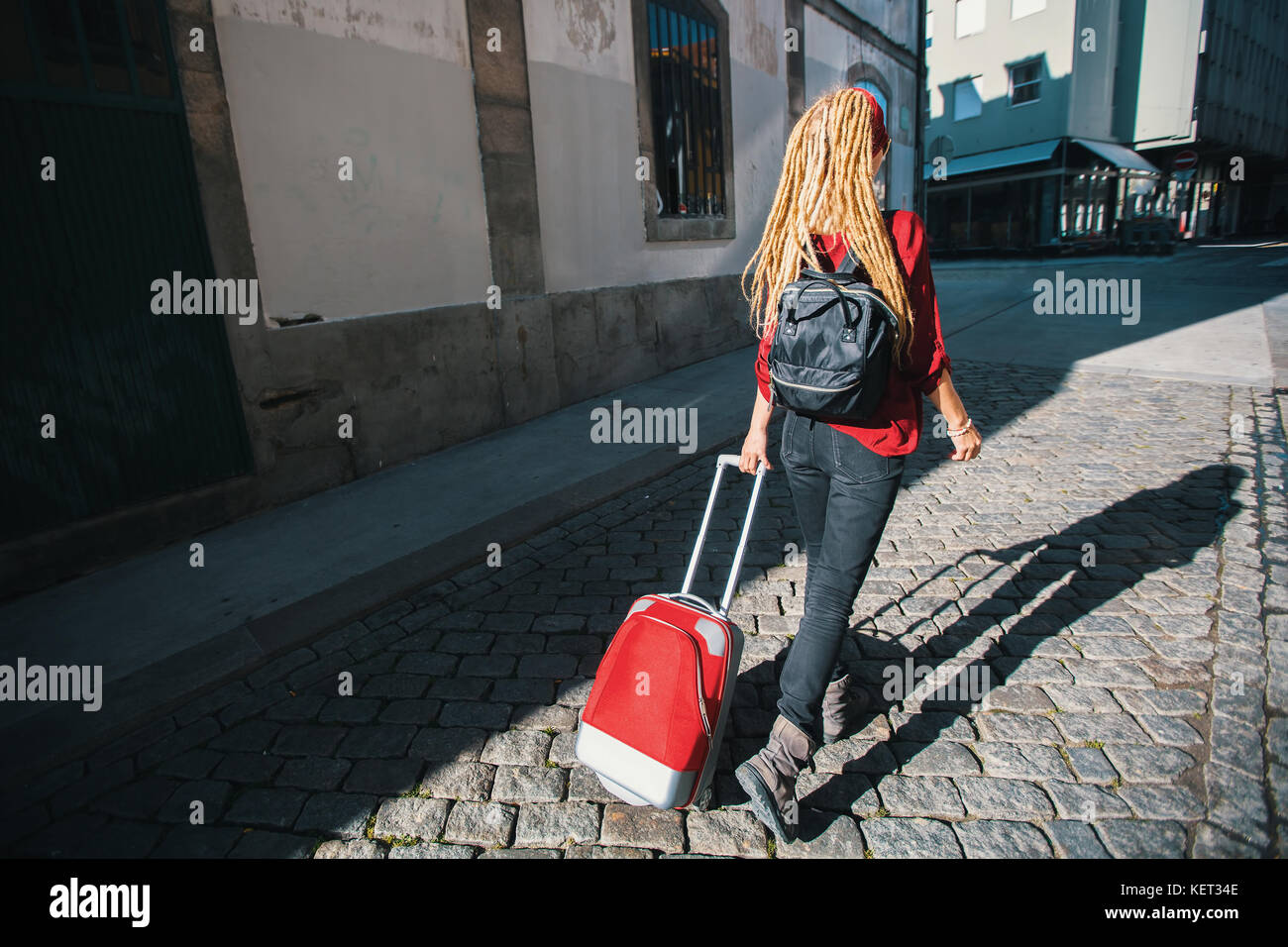 Junge Frau auf dem Bürgersteig mit einem roten Koffer. Rückansicht. Reise und Tourismus. Stockfoto