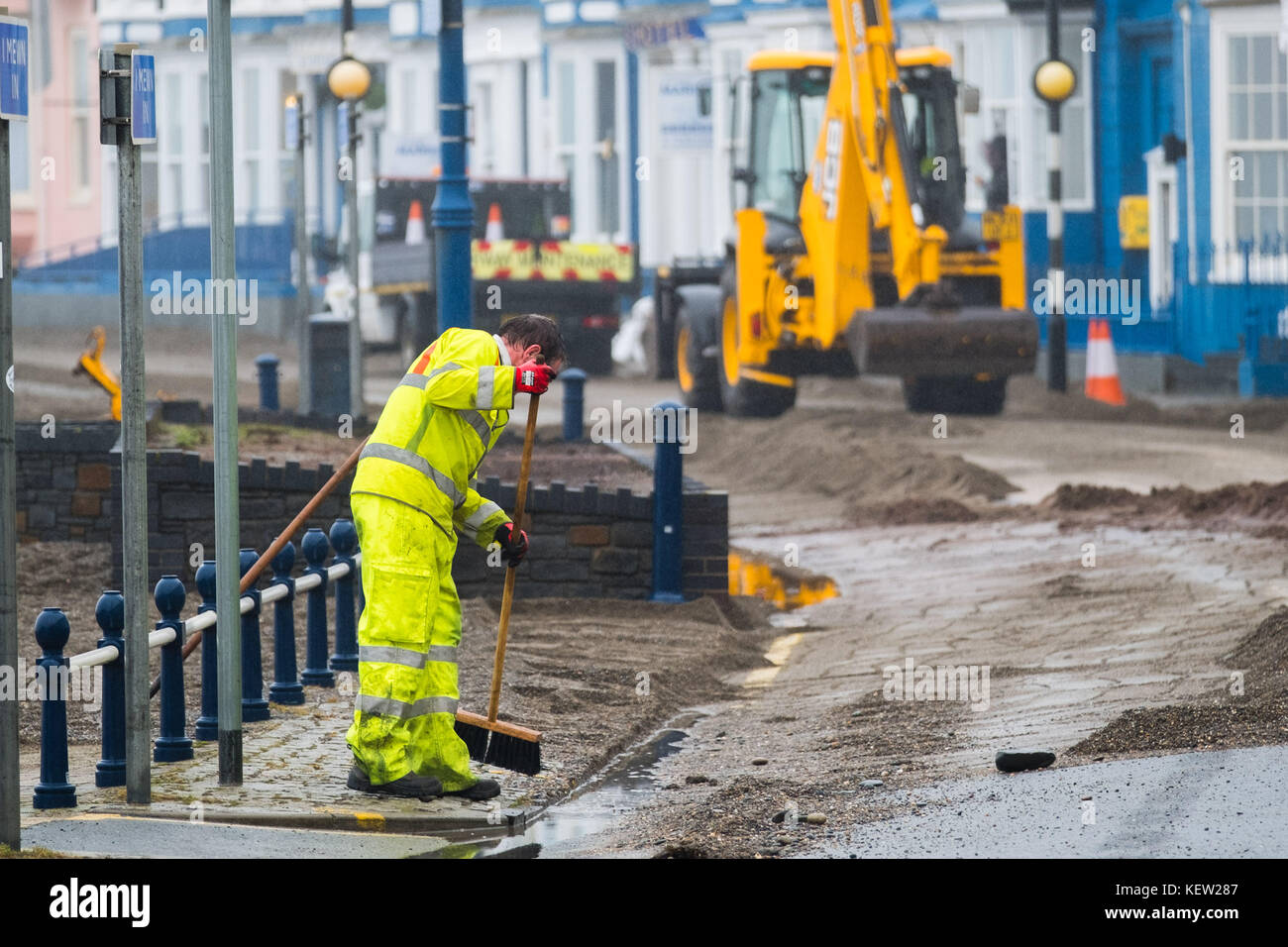 Aberystwyth Wales Vereinigtes Königreich, Montag, 23. Oktober 2017 Vereinigtes Königreich Wetter: Nach dem Sturm Wind und wilde Meere des Sturms Brian, Mitarbeiter des gemeinderats und Auftragnehmer, die JCB's und Bürsten verwenden, beginnen mit der Reinigung der Hunderte Tonnen von Sandsand und Kies, die an der Promenade und am Meer in Aberystwyth an der Küste von Cardigan Bay in westwales angespült wurden. Sie erwarten, dass die Straße bis zum Nachmittag wieder für den Verkehr freigegeben wird. Foto: Keith Morris/Alamy Live News Stockfoto