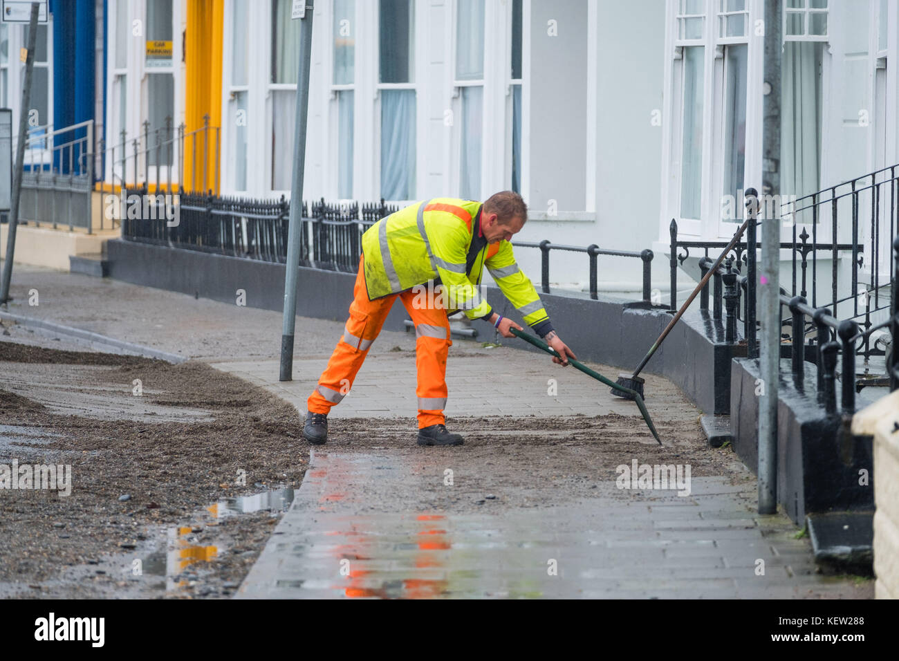 Aberystwyth Wales Vereinigtes Königreich, Montag, 23. Oktober 2017 Vereinigtes Königreich Wetter: Nach dem Sturm Wind und wilde Meere des Sturms Brian, Mitarbeiter des gemeinderats und Auftragnehmer, die JCB's und Bürsten verwenden, beginnen mit der Reinigung der Hunderte Tonnen von Sandsand und Kies, die an der Promenade und am Meer in Aberystwyth an der Küste von Cardigan Bay in westwales angespült wurden. Sie erwarten, dass die Straße bis zum Nachmittag wieder für den Verkehr freigegeben wird. Foto: Keith Morris/Alamy Live News Stockfoto