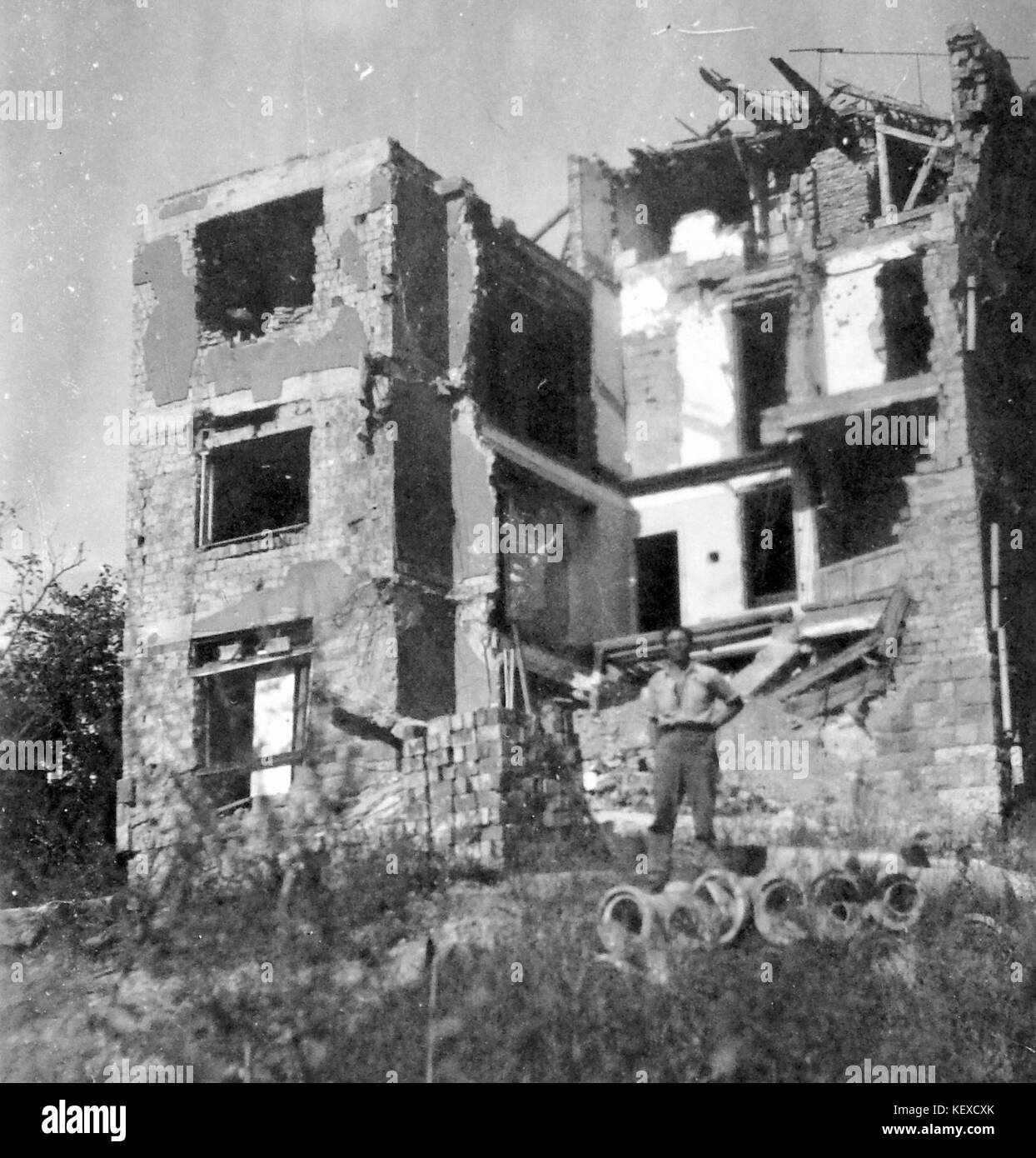 Ein Schwarzweiß-Foto, das ein schwer beschädigtes mehrstöckiges Gebäude darstellt, wahrscheinlich aus Kriegszerstörungen, mit einem Mann, der vor den Ruinen steht. Stockfoto