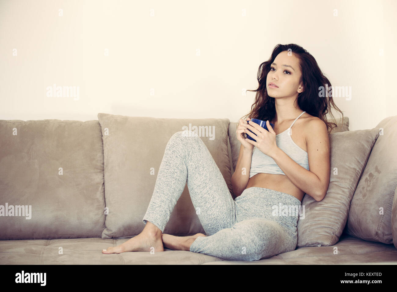 Junge hübsche asiatische Frau sitzt entspannt und nachdenklich auf der Couch nachdenklichen Gesichtsausdruck blau Tasse in den Händen Stockfoto