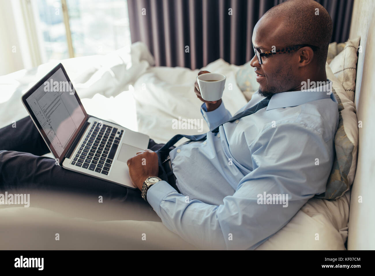 Mann in formeller Kleidung, der am Laptop arbeitet, während er im Bett liegt. Geschäftsmann, der an einem Laptop arbeitet, während er Kaffee in seinem Hotelzimmer trinkt. Stockfoto