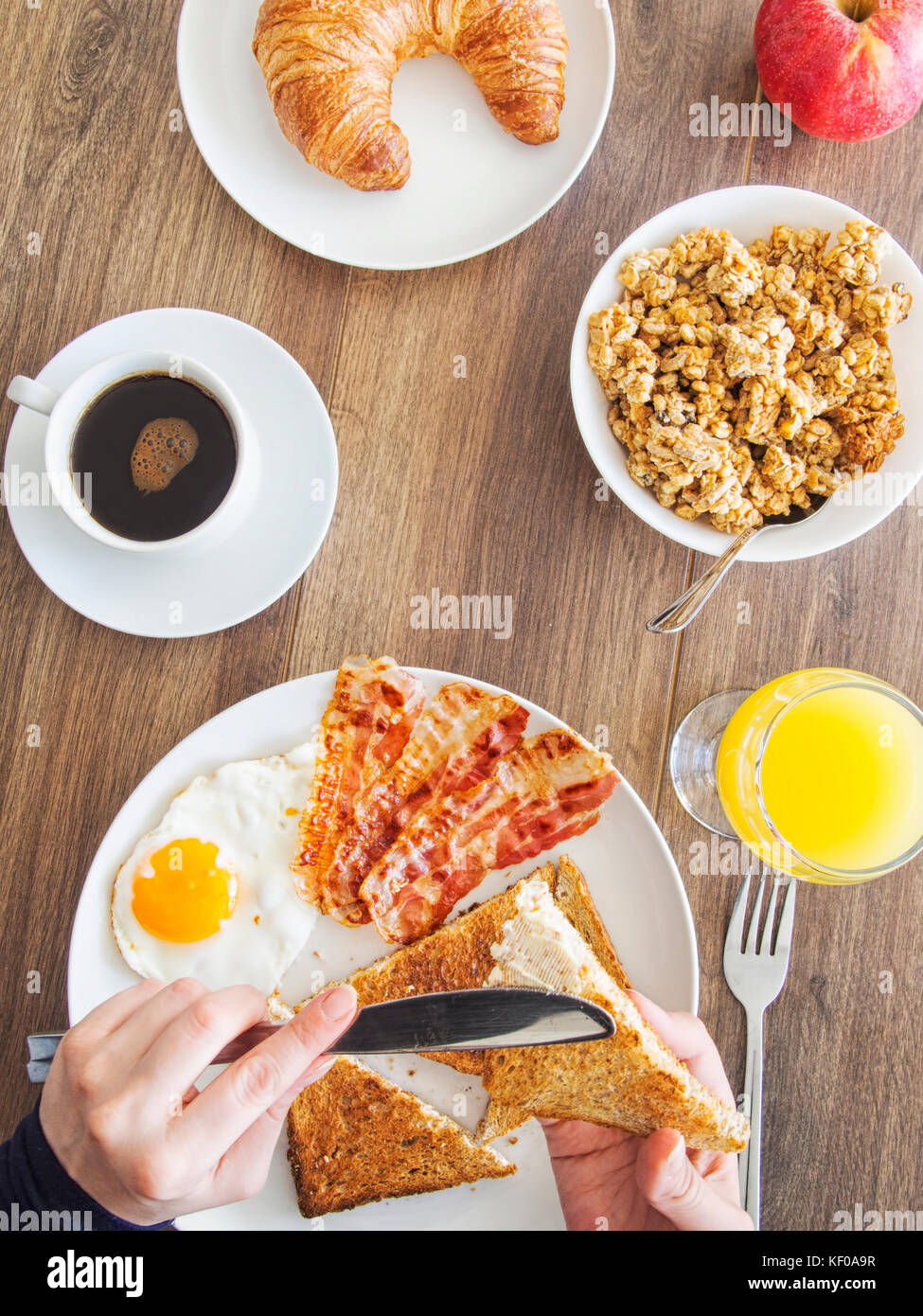 Frühstück mit Eiern, Speck, Kaffee, Croissants, Müsli, Saft und Apple. Frauen Verbreitung Butter auf Toast. Stockfoto