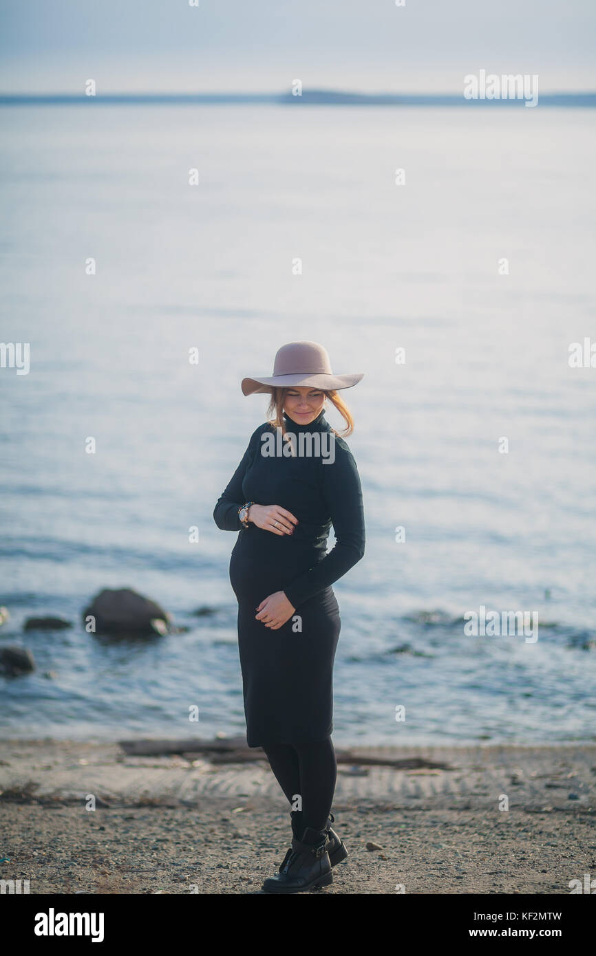 Schöne junge schwangere Frau in einem schwarzen Rollkragenpullover und Schwarz, enger Rock, Klassik beige Hut vor einem blauen Meer im Herbst Tag posing Stockfoto