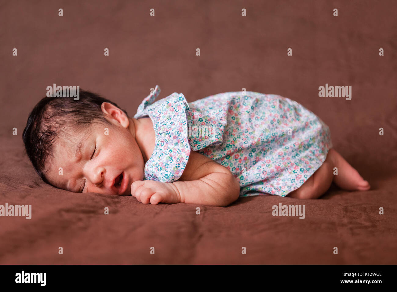 Cute zwei Wochen alte Neugeborene Mädchen mit einem geblümten Kleid, friedlich schlafend im Bett in der fetalen Position/neugeborenes Baby süße Mädchen schlafen schlafen Stockfoto
