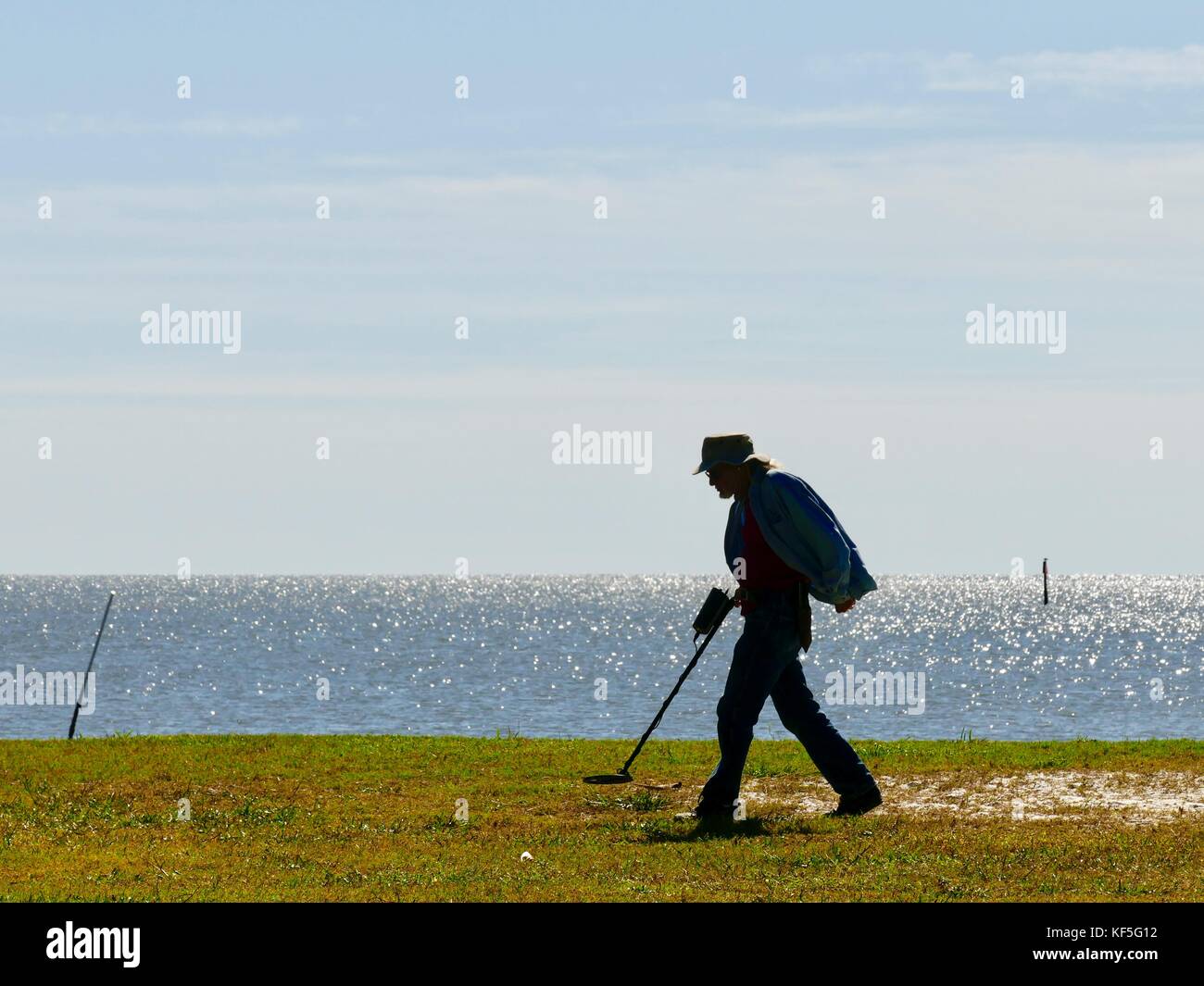 Mann in silhouette Suche mit einem Metalldetektor im City Park mit Golf Wasser im Hintergrund. Cedar Key, Florida, USA Stockfoto