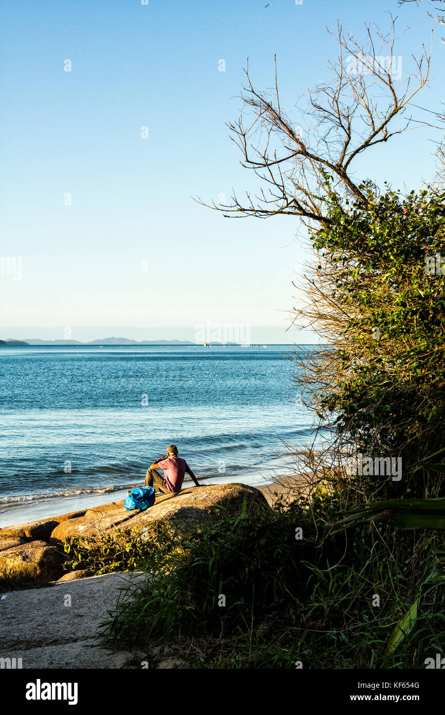 Rückansicht eines Mannes, der auf einem Felsen am Daniela Beach sitzt. Florianopolis, Santa Catarina, Brasilien. Stockfoto