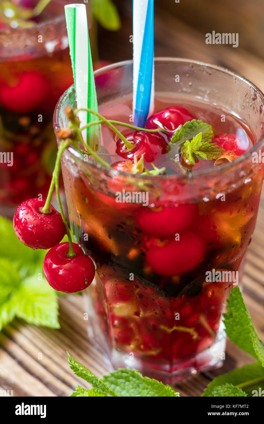 Gläser erfrischenden Drink gewürzt mit frischem Obst und mit Kirschen mit Tautropfen bedeckt gehalten. Holz- Hintergrund Stockfoto