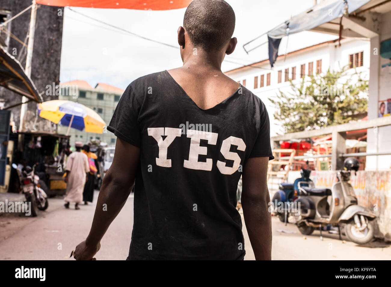 Afrikanischer Mann, der auf einer Marktstraße läuft, fotografiert von hinten, mit einem schwarzen T-Shirt, auf dem sich JA in weißen Großbuchstaben auf dem Rücken befindet. Stockfoto