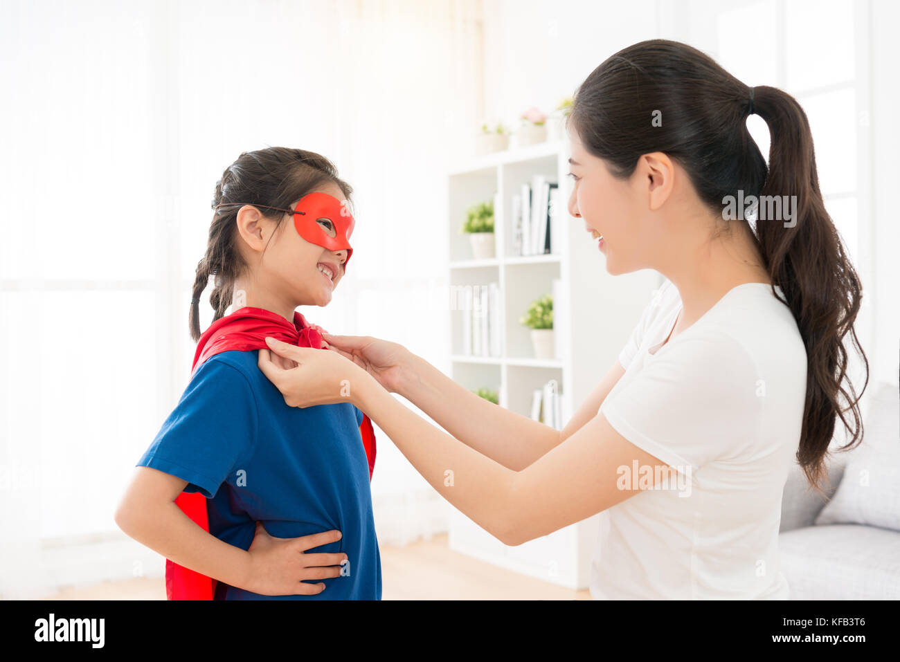 Gerne attraktive Mutter helfen fröhliche kleine süße Tochter spielen als Superheld zu Hause mit rotem Umhang und Maske während Urlaub. Stockfoto