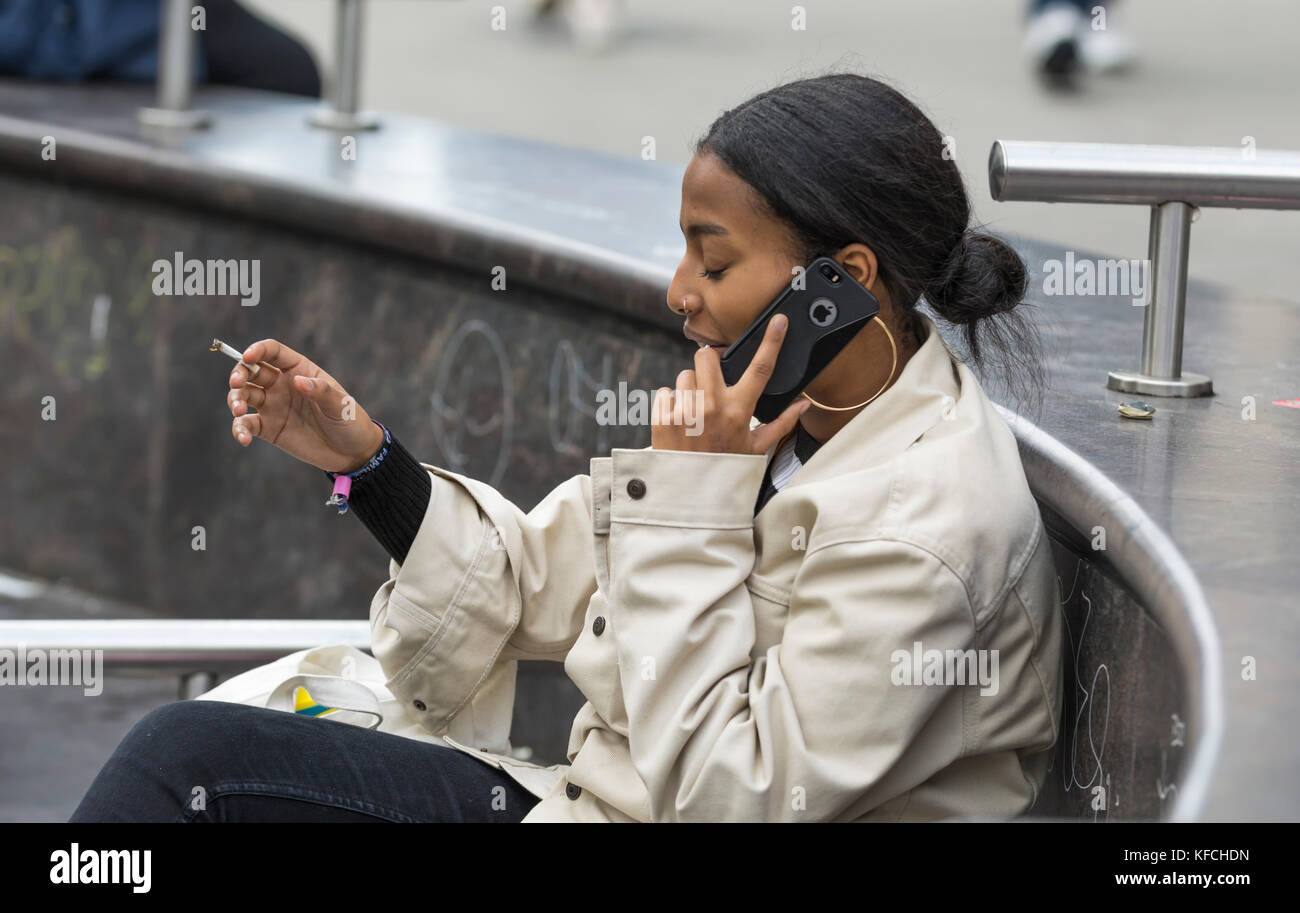 Junge Frau, die das Telefon benutzt, während sie in Großbritannien eine Zigarette raucht. Stockfoto