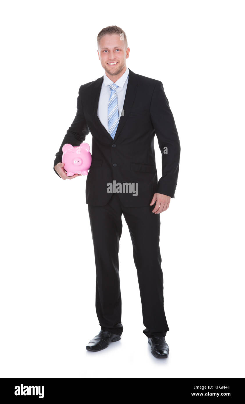 Stilvolle Geschäftsmann im Anzug steht ein rosa Keramik Sparschwein unter den Arm, wie er lächelt in die Kamera in einem konzeptionellen finanzielle Einsparungen m Stockfoto