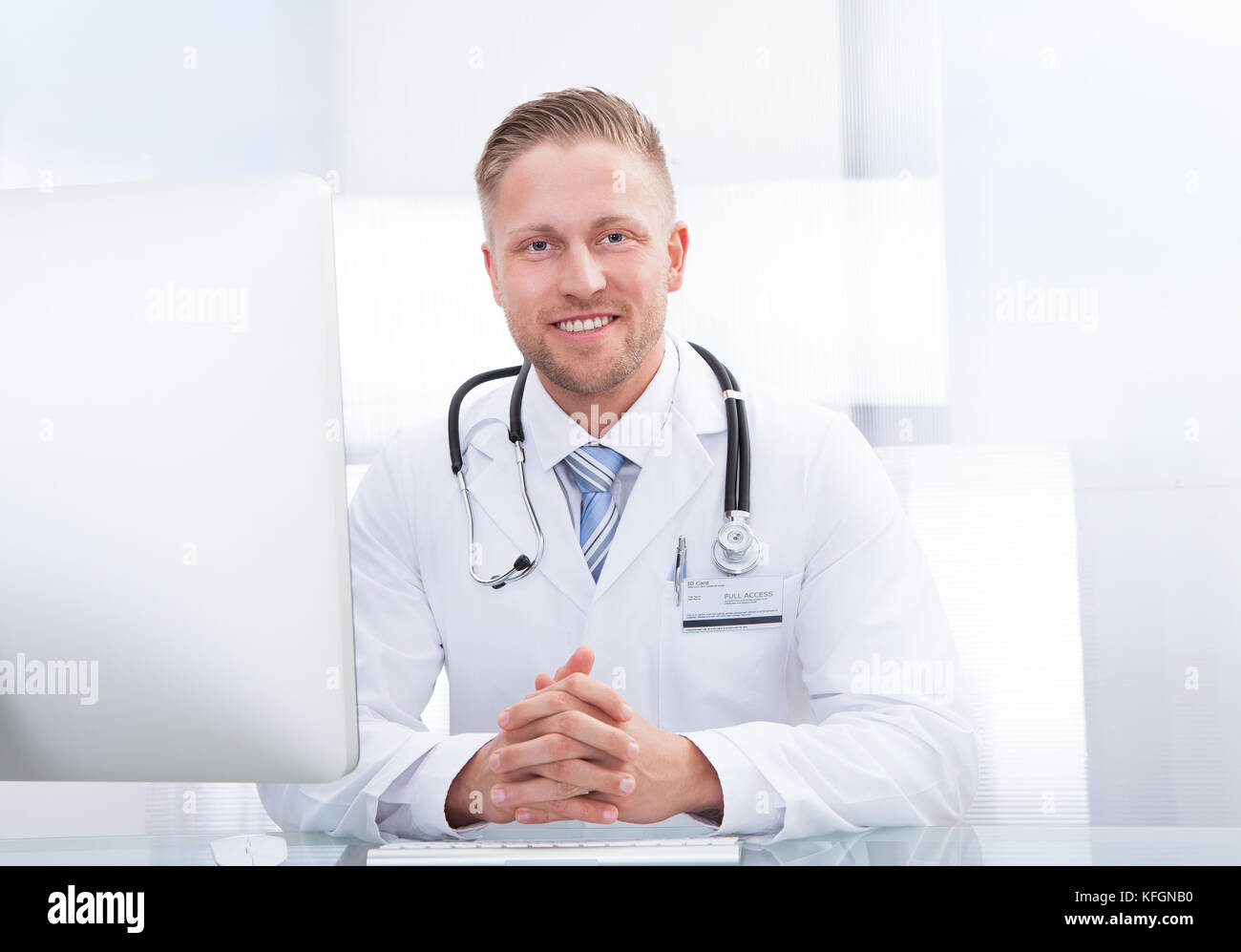 Lächelnd Arzt oder Berater an einem Schreibtisch mit seinem Stethoskop um den Hals in die Kamera schaut Sitzen Stockfoto