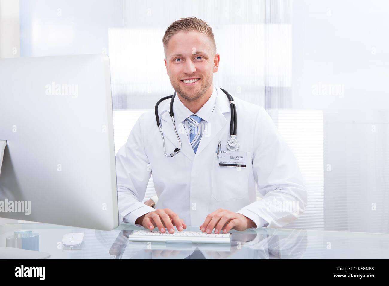 Lächelnd Arzt oder Berater an einem Schreibtisch mit seinem Stethoskop um den Hals in die Kamera schaut Sitzen Stockfoto